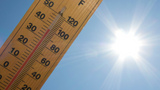 A wooden thermometer showing high temperatures in front of clear blue sky and the sun radiating brilliant light.